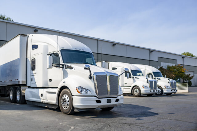 Row of the big rig semi trucks tractors with dry van semi trailers standing in warehouse docks loading cargo for the next delivery freight